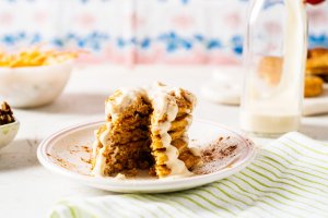 Mini Carrot Cake Pancakes with Vanilla Greek Yogurt Frosting from Holley Grainger - Image of five cakes stacked on a white plate with icing drizzle and a wedge shaped piece cut through the stack