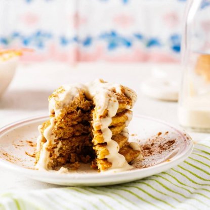 Mini Carrot Cake Pancakes with Vanilla Greek Yogurt Frosting from Holley Grainger - Image of five cakes stacked on a white plate with icing drizzle and a wedge shaped piece cut through the stack